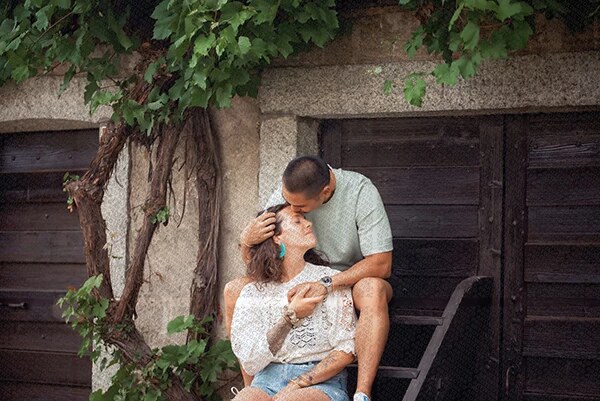 Couple assis devant une porte en bois, l'homme embrasse tendrement le front de la femme entourée de verdure