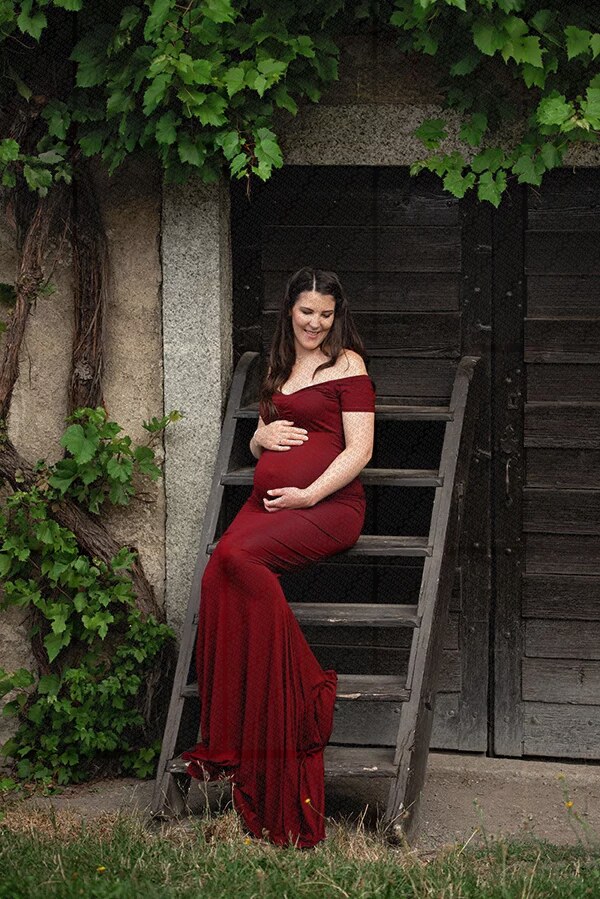 Femme enceinte souriante en robe longue rouge assise sur un escalier en bois devant un mur végétalisé