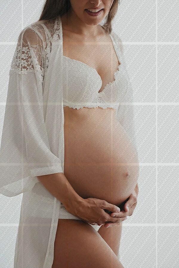 Femme enceinte en lingerie blanche et peignoir léger tenant son ventre arrondi avec douceur