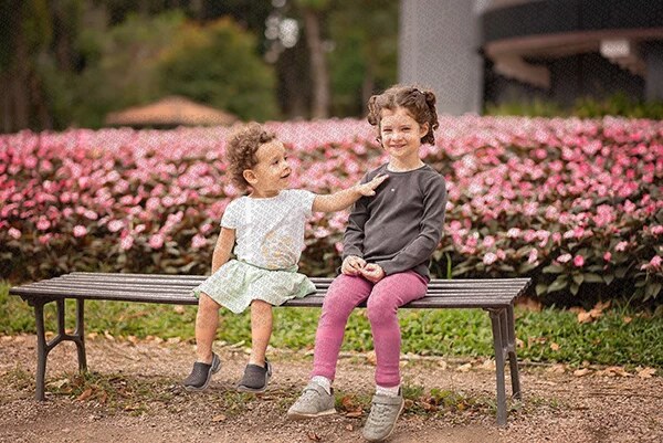 Deux enfants assis sur un banc dans un parc fleuri, l'un souriant avec la main posée sur l'épaule de l'autre