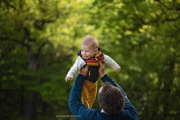 Père soulevant son bébé souriant dans un parc avec un fond flou d'arbres verts