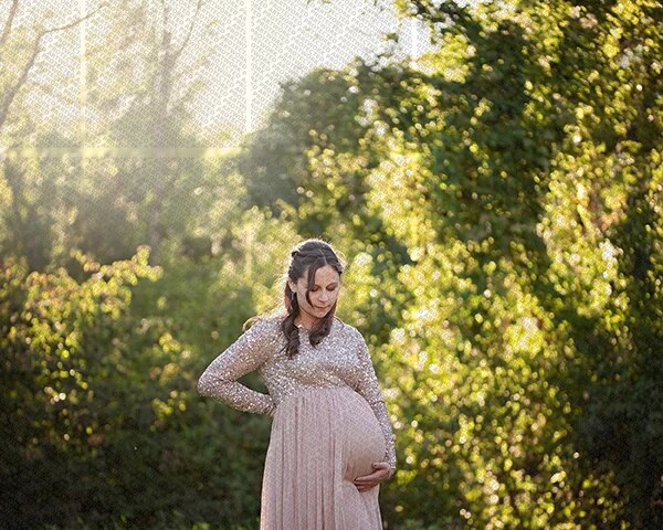 Femme enceinte en robe longue scintillante, caressant son ventre dans un jardin lumineux au coucher du soleil