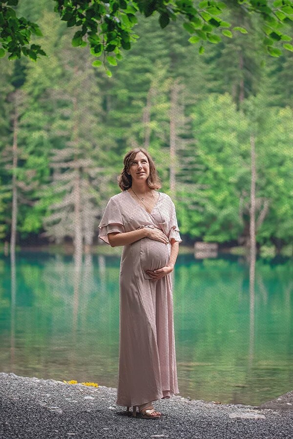 Femme enceinte en robe rose clair posant au bord d'un lac entouré d'arbres verts en arrière-plan
