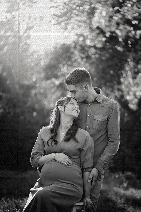 Couple en séance photo grossesse en extérieur avec le futur père embrassant la tête de la femme enceinte assise