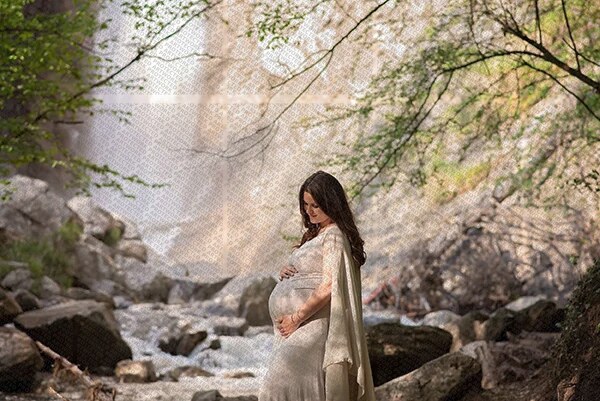 femme enceinte en robe claire debout près d'un ruisseau dans un paysage naturel rocheux et boisé