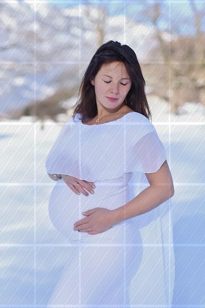 Femme enceinte en robe blanche posant dans un paysage enneigé, les mains sur le ventre, regard baissé