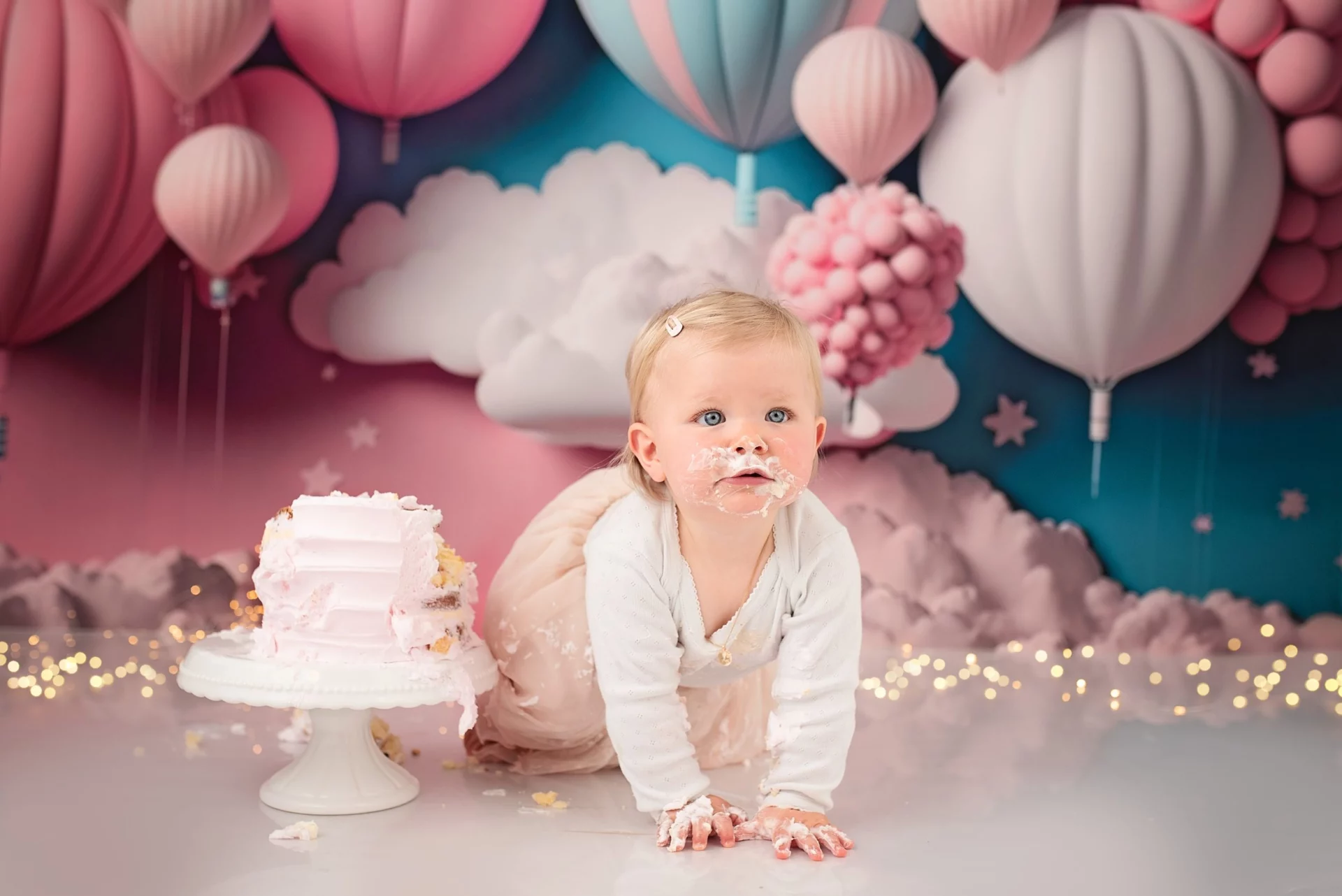 Bébé avec de la crème sur le visage, accrochant au sol près d'un gâteau rose partiellement mangé, décor ballon pastel