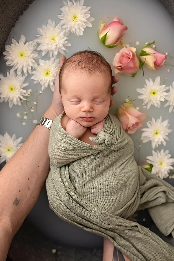 Nouveau-né endormi enveloppé dans un tissu vert, tenu par une main, entouré de fleurs blanches et roses