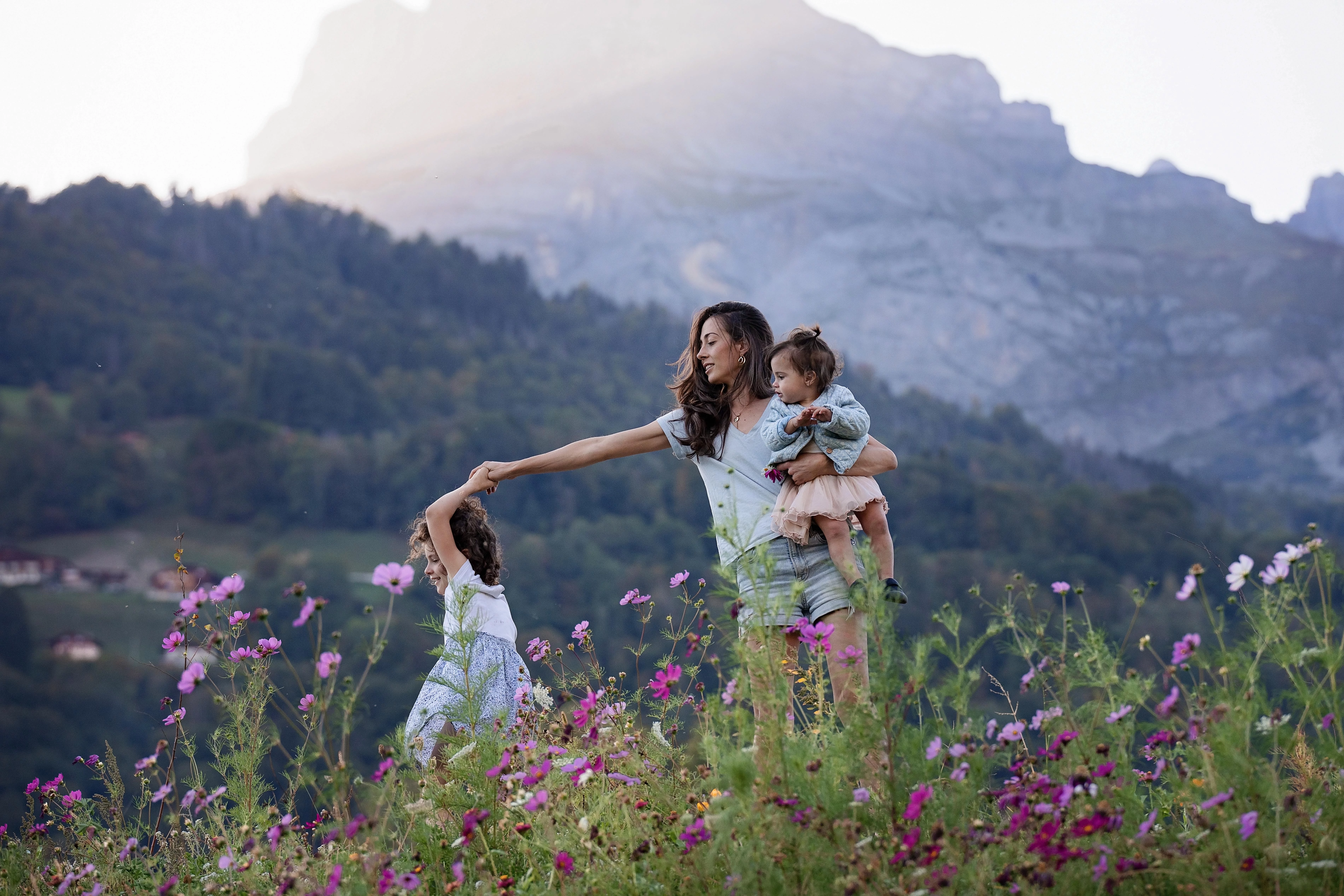 Mère tenant un enfant dans un champ de fleurs sauvages, jouant avec une autre enfant avec montagnes en arrière-plan